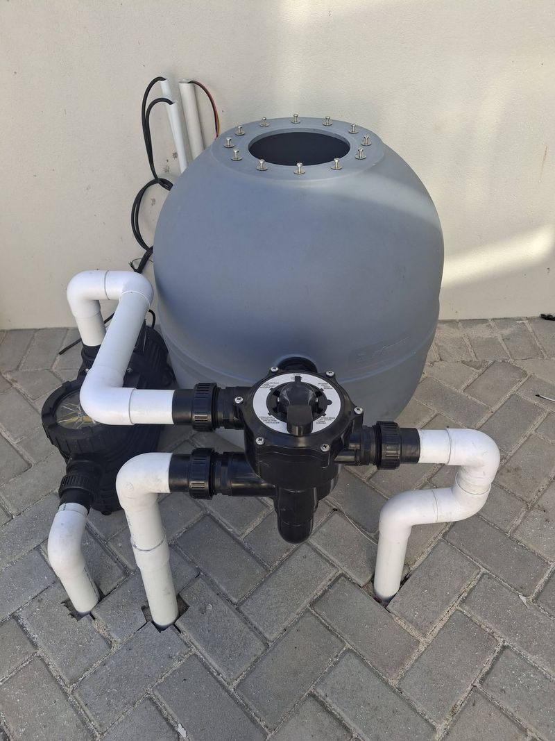 A grey sand filter with a black multiport valve and pool pump, connected with white PVC pipework on a paved pad.